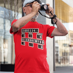 A man wearing a bright red t-shirt with a black and white scoreboard graphic, a gray cap, and glasses looking through binoculars outdoors.