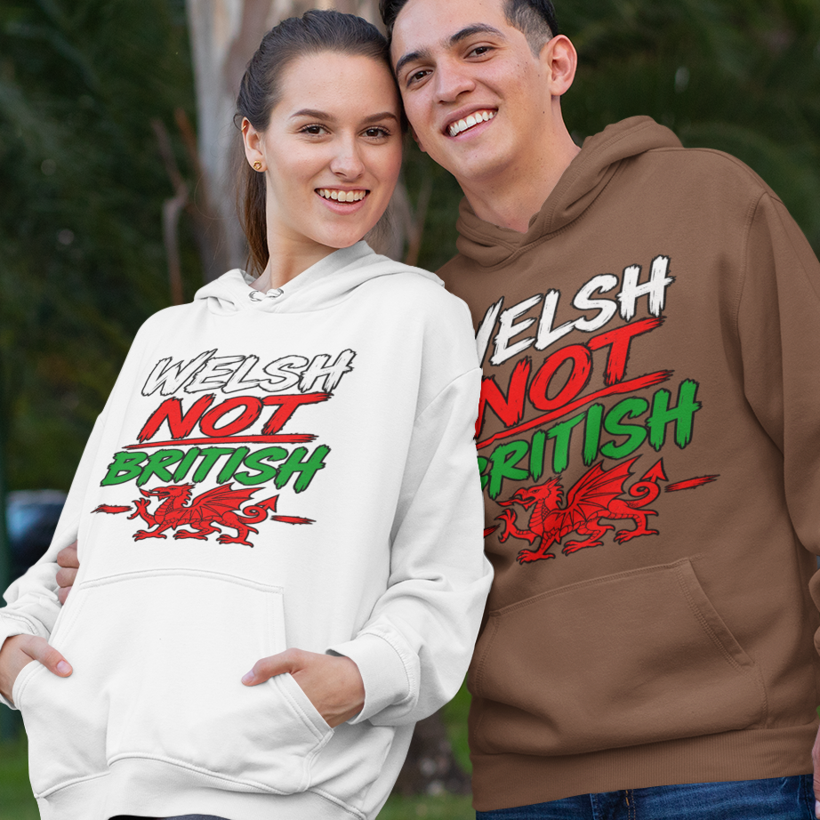 Two people standing outdoors in front of trees, each wearing a hoodie with the slogan 'Welsh Not British' and a red dragon graphic. The person on the right wears a brown hoodie, and the person on the left wears a white hoodie.