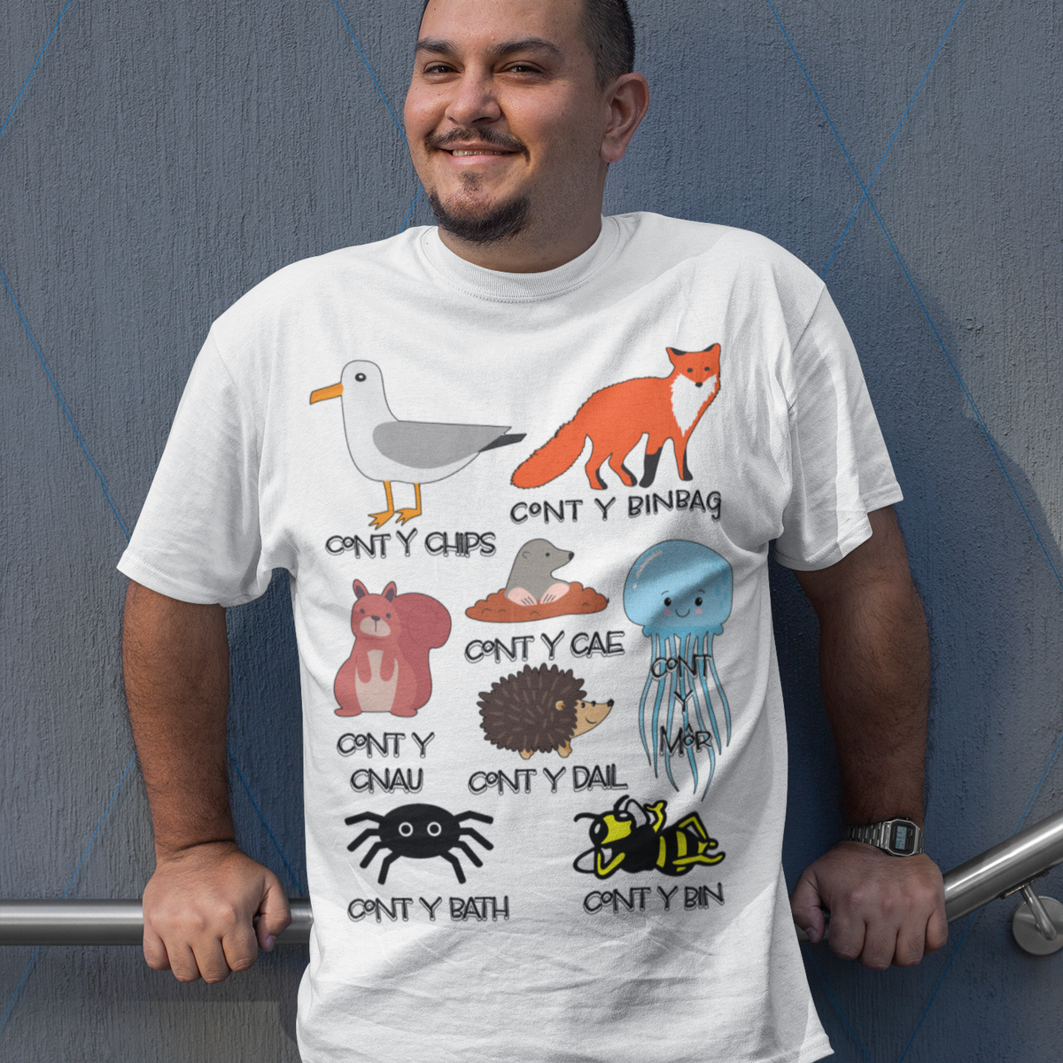 A man wearing a white graphic t-shirt with various Welsh animal illustrations and text, standing against a gray textured wall.