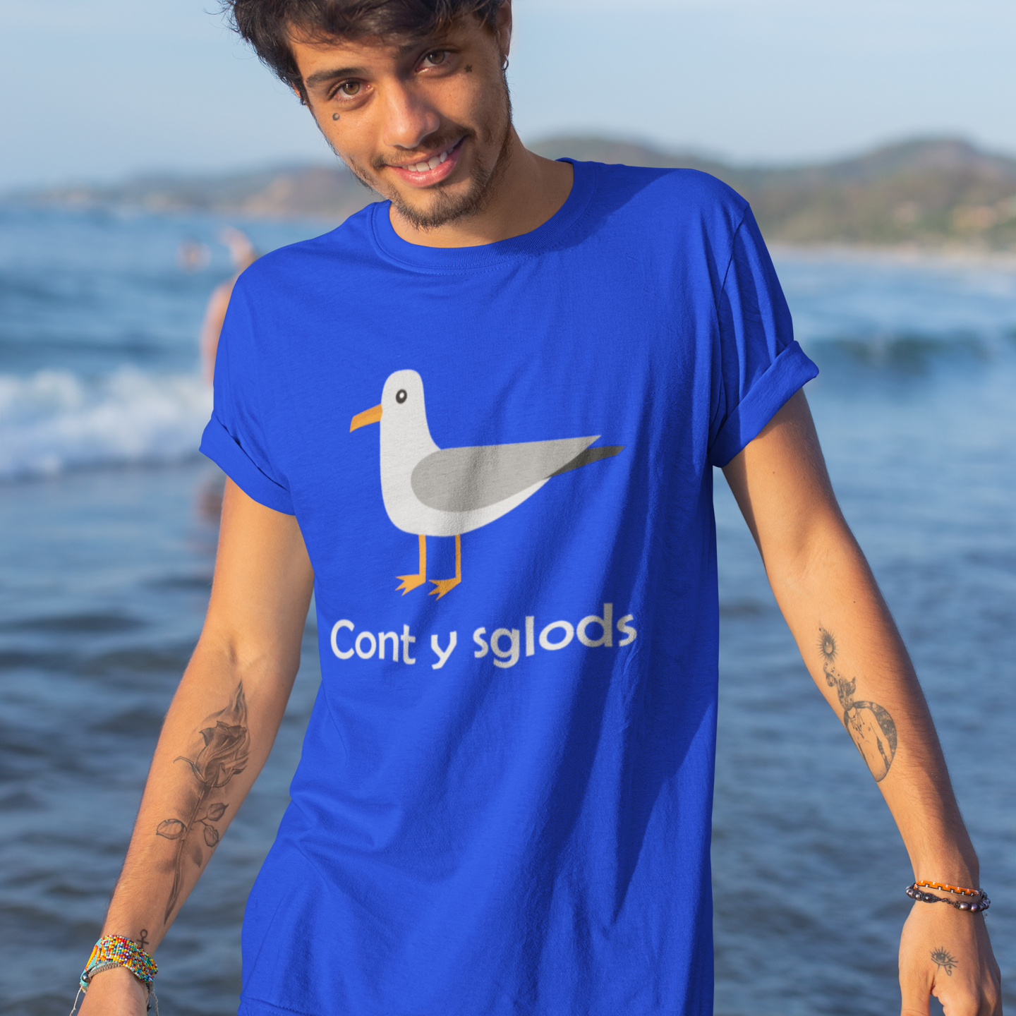 A man wearing a bright blue graphic t-shirt with a seagull illustration and the text 'Cont y sglods' standing in shallow ocean water with a beach background.