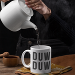 A person pouring hot water from a white ceramic teapot into a white ceramic mug printed with 'DUW DUW' in gray. The mug sits on a patterned napkin on a rustic wooden table, with a wooden pestle and mortar in the background.