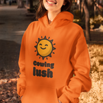 A woman wearing a bright orange hoodie with a yellow smiling sun graphic and the text 'cowing lush' printed on the front, standing outdoors in a sunlit park setting.