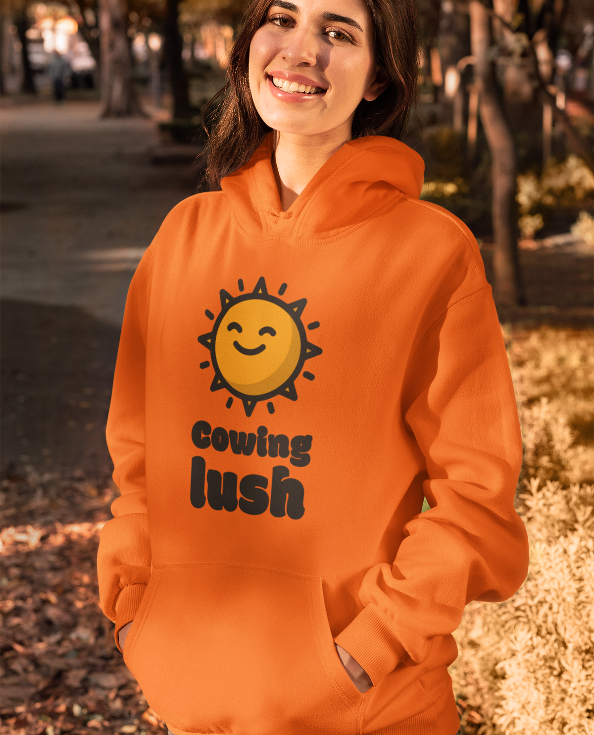 A woman wearing a bright orange hoodie with a yellow smiling sun graphic and the text 'cowing lush' printed on the front, standing outdoors in a sunlit park setting.