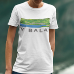 A person wearing a white short-sleeve graphic T-shirt with a landscape print and the text 'Y BALA' on the front, paired with blue jeans, standing outdoors against a blurred mountain and lake background.