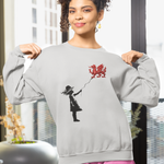 A woman modeling a light gray crewneck sweatshirt with a black and red graphic of a girl flying a red dragon balloon, paired with pink shorts in a modern, sunlit room.
