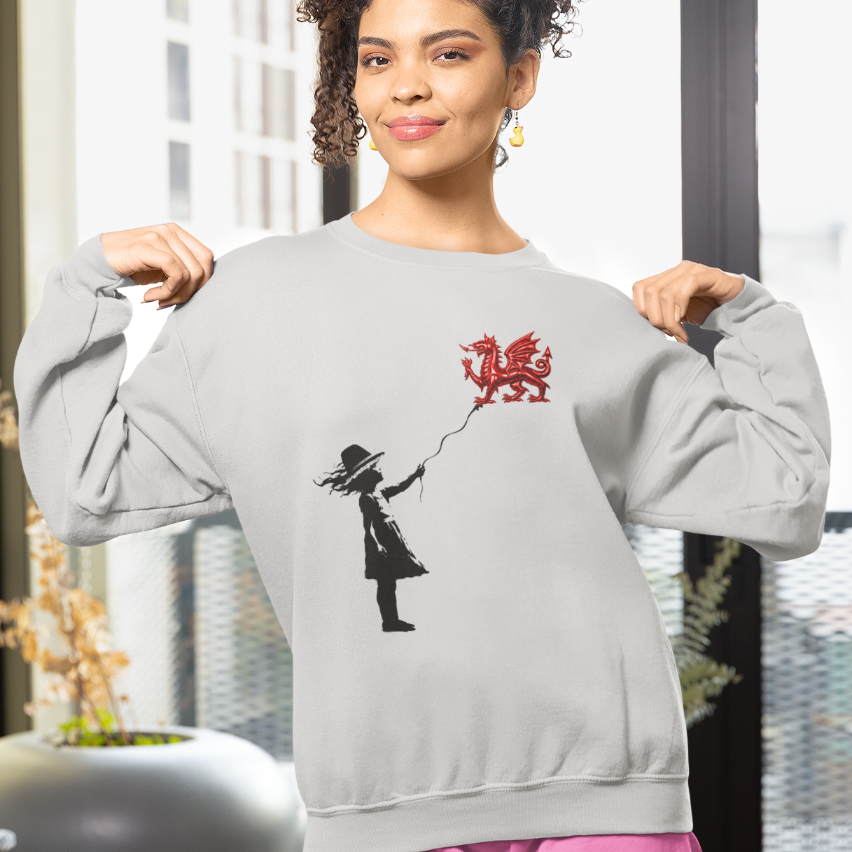 A woman modeling a light gray crewneck sweatshirt with a black and red graphic of a girl flying a red dragon balloon, paired with pink shorts in a modern, sunlit room.