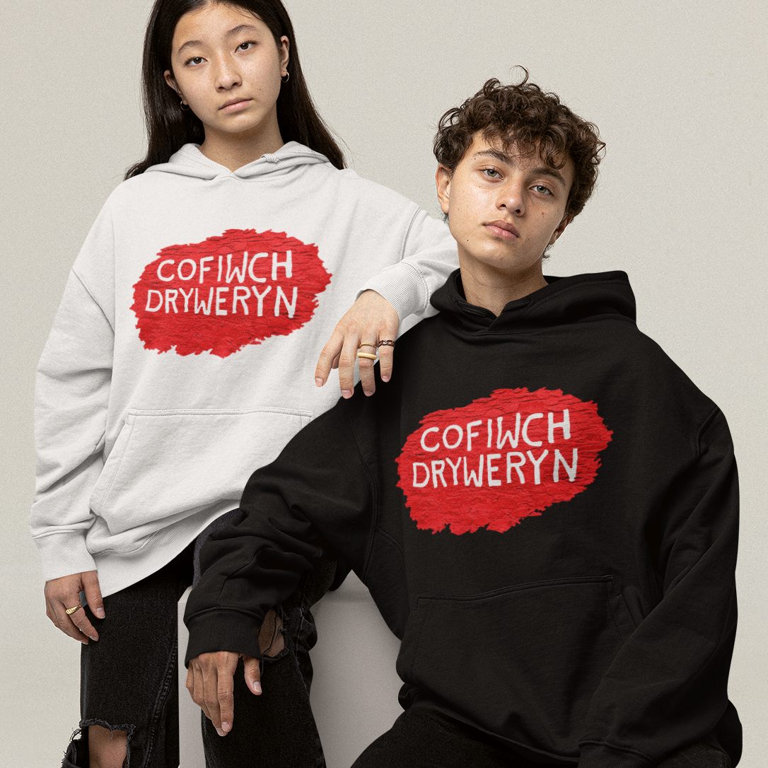 Two models posing in matching oversized hoodies, one white and one black, both featuring a red paint-splatter style graphic with the text 'COFIWCH DRYWERYN' in white. The models are styled with distressed black jeans and white sneakers against a neutral studio backdrop.