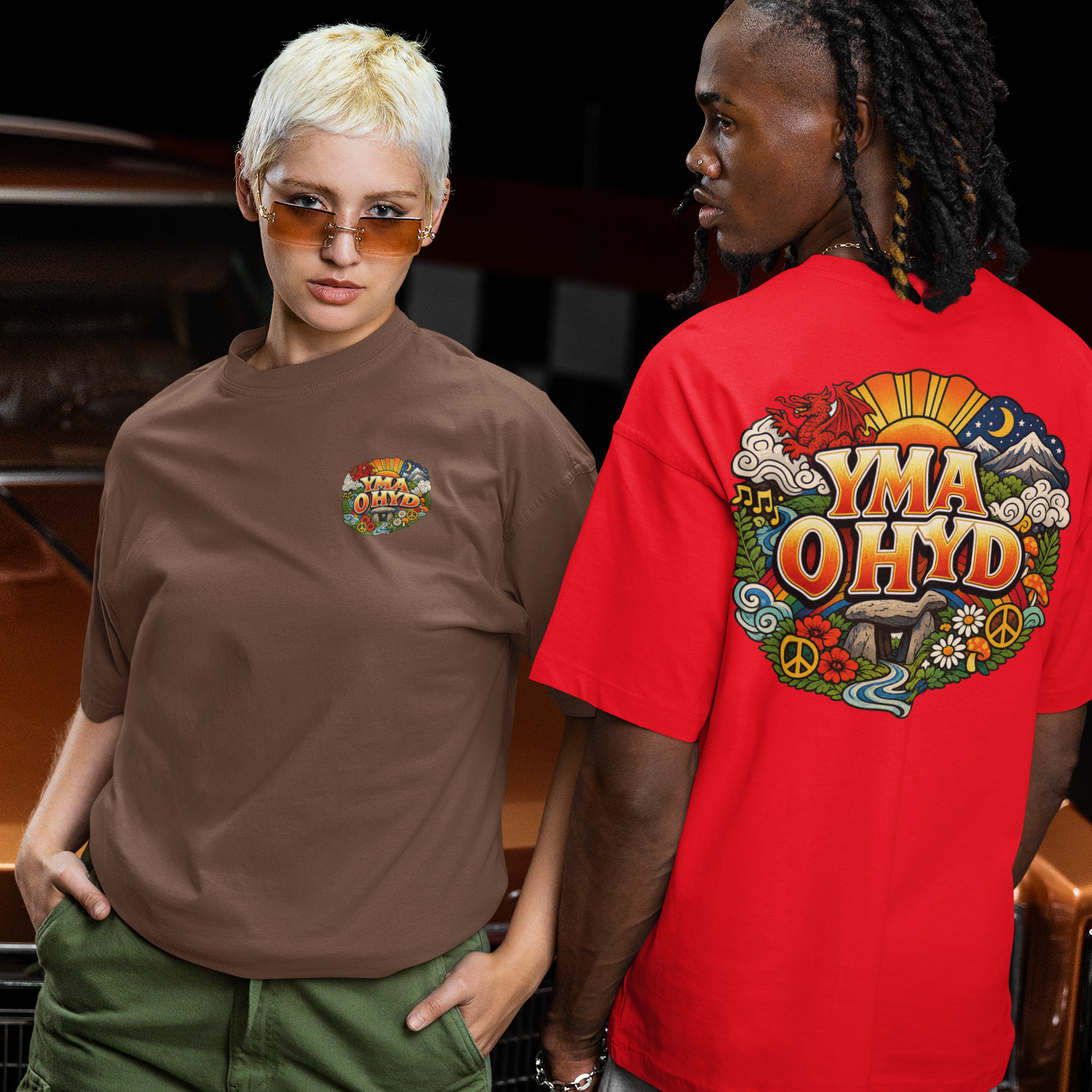 Two people standing next to a classic car, one wearing a brown t-shirt with a colorful design and the other wearing a red t-shirt with 'YMA OHYD' text.