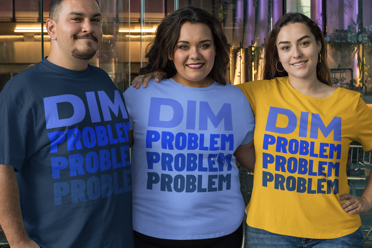 Three women standing together in an industrial interior wearing graphic t-shirts with the text 'DIM PROBLEM PROBLEM PROBLEM' in blue. The shirt colors are navy blue, light blue, and yellow.