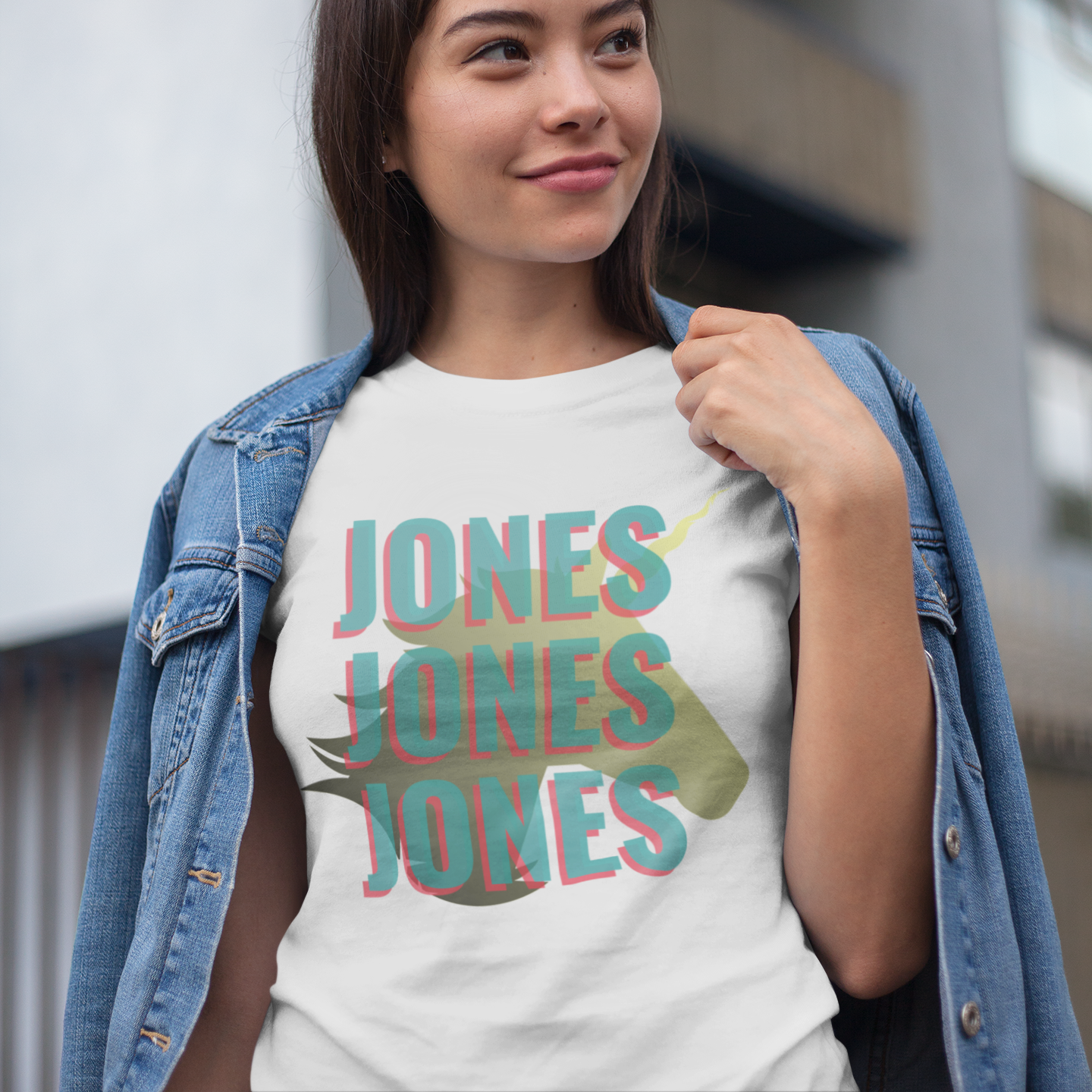 A woman wearing a white graphic t-shirt with 'JONES' printed in a 3D-effect teal and pink font over a green leaf, styled with an open blue denim jacket in an outdoor setting.
