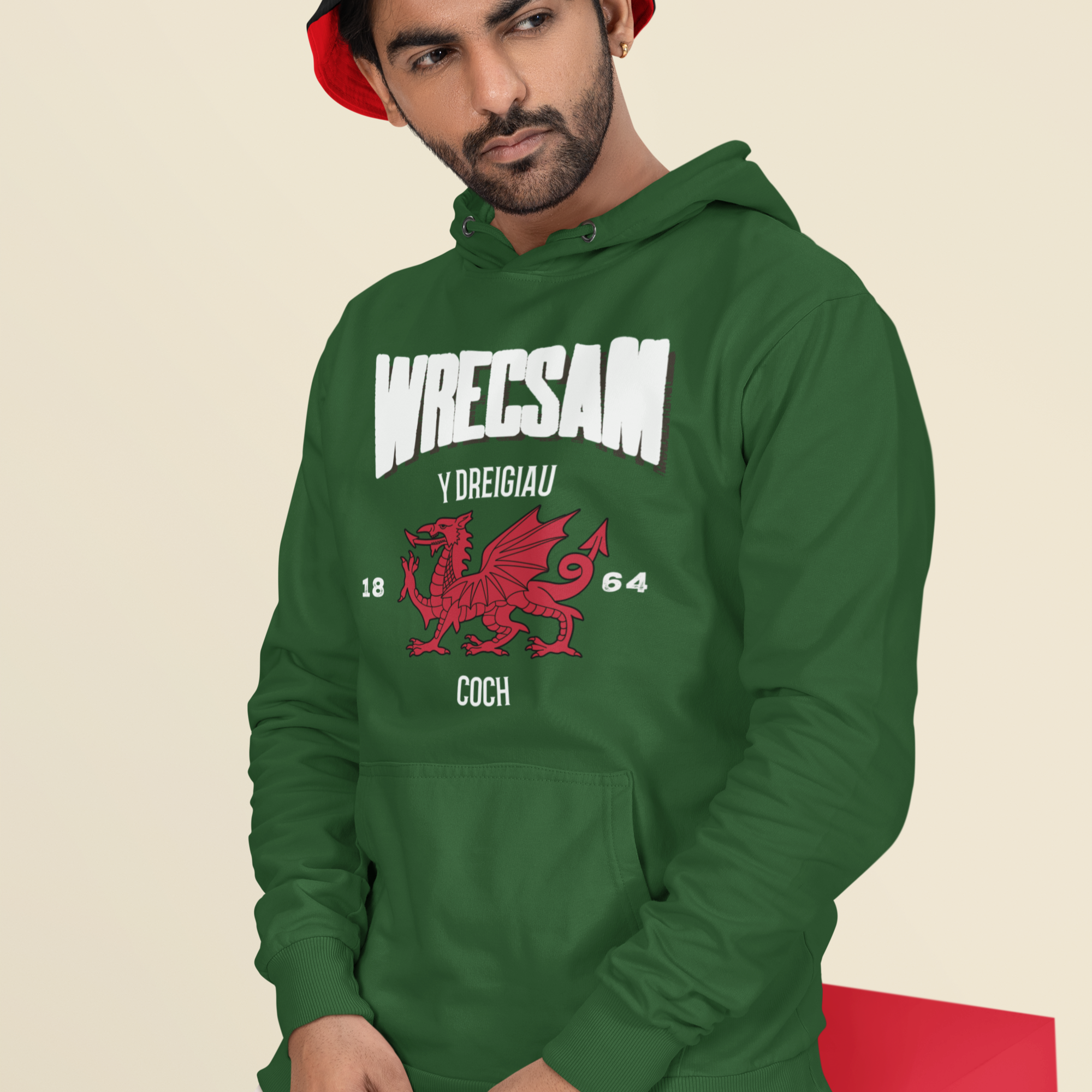 Man wearing a green hoodie with Wrecsam and graphics, sitting on a red box against a beige background