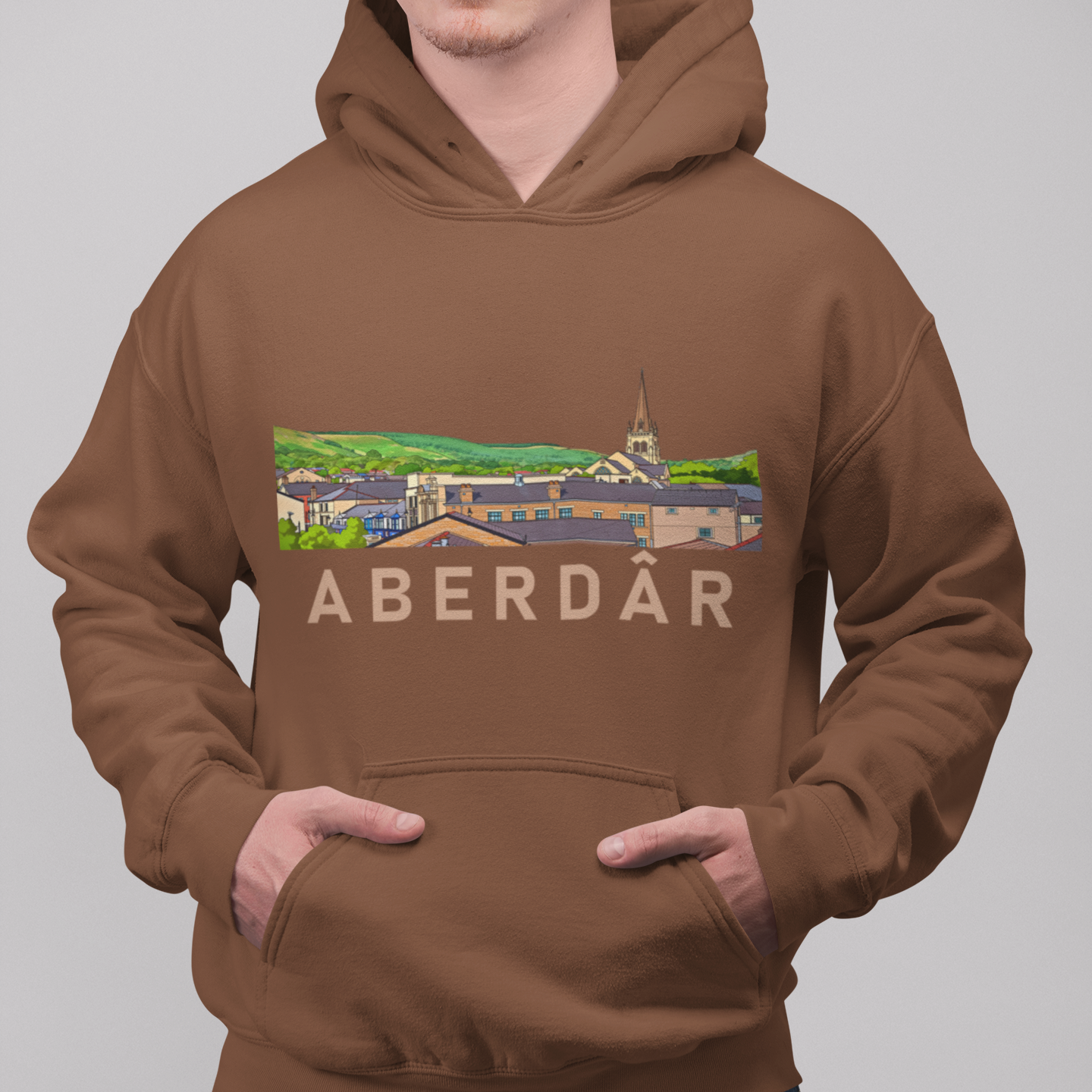 A man wearing a brown pullover hoodie with a colorful graphic of a town and the text 'ABERDÁR' across the chest, paired with blue jeans, against a solid gray background.