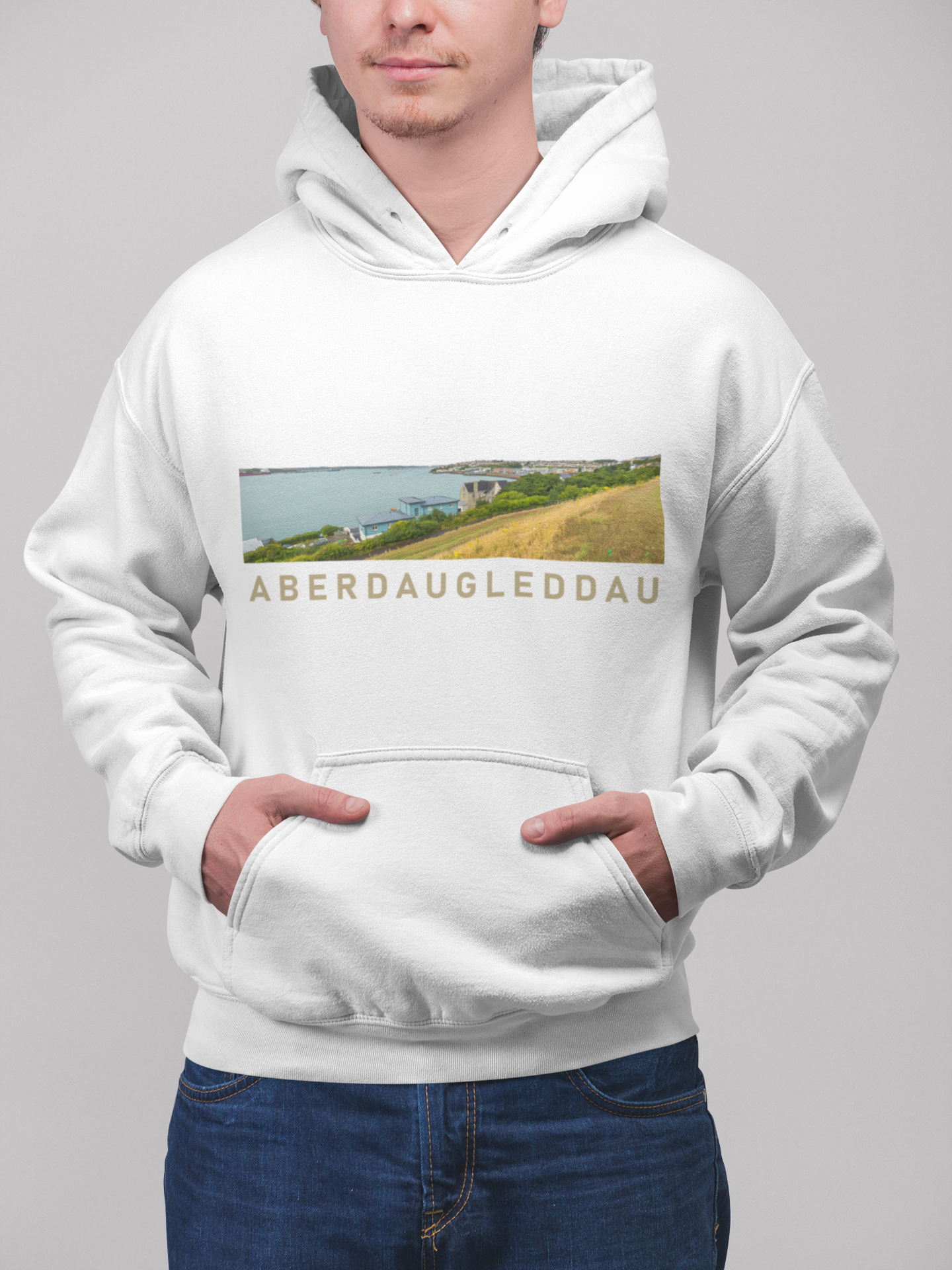 A man wearing a white pullover hoodie with a rectangular landscape graphic on the chest and the text 'ABERDAUGLEDDAU' printed below it, paired with blue jeans against a plain gray studio background.