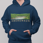 A man wearing a navy blue hoodie with a rectangular scenic graphic on the chest that reads 'ABERGWAUN' in beige letters, paired with blue jeans against a plain gray studio background.