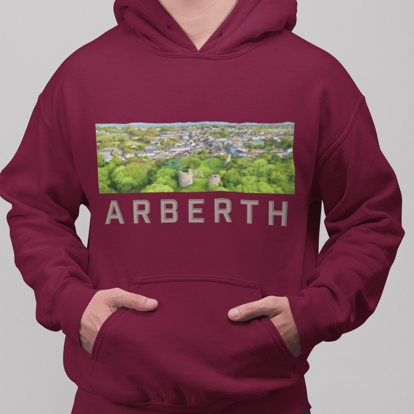 A man wearing a maroon pullover hoodie with a graphic print of a town and the word 'ARBERTH' on the front, paired with blue jeans, against a solid gray background.