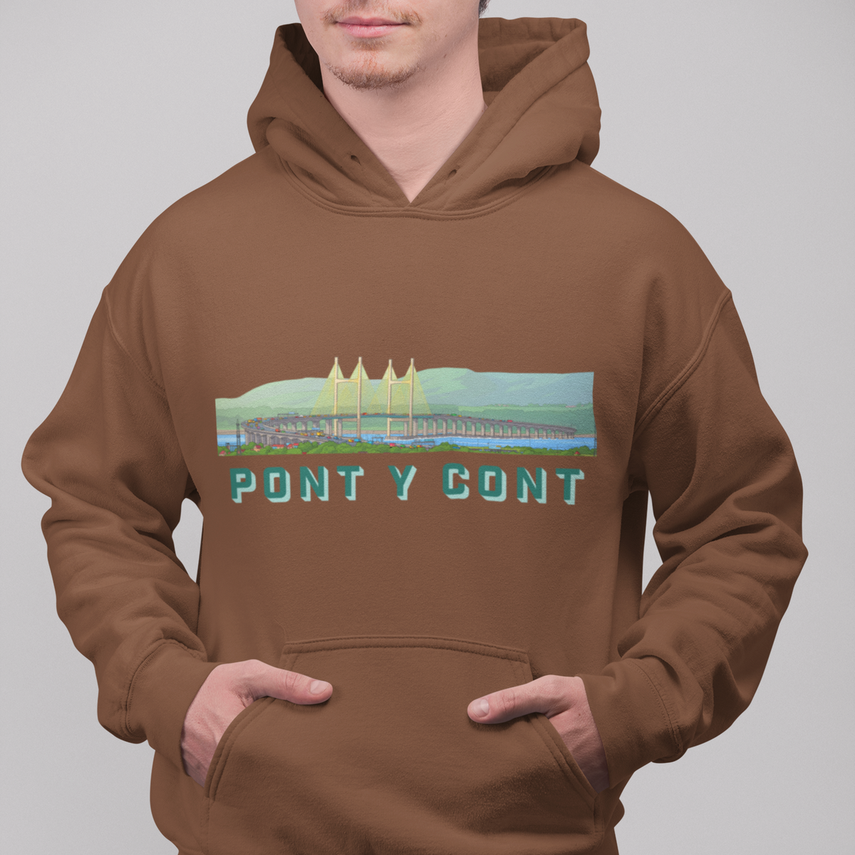 A man wearing a brown pullover hoodie with a colorful graphic of the Pont Y Cont bridge and the text 'PONT Y CONT' printed on the chest, paired with blue jeans against a solid gray background.