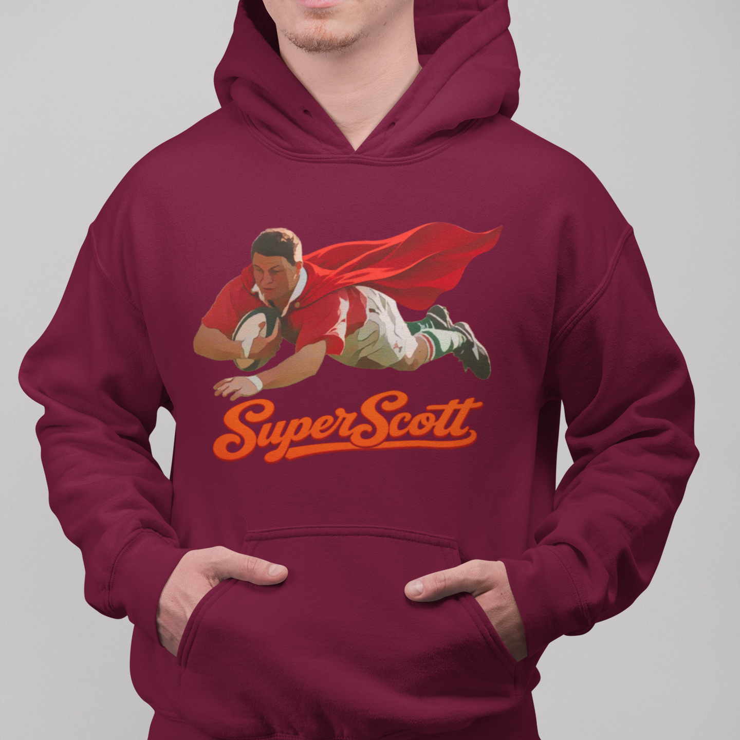 A man wearing a maroon hoodie with a graphic of a rugby player in a red cape and the text 'Super Scott' in orange, paired with blue jeans against a solid gray background.