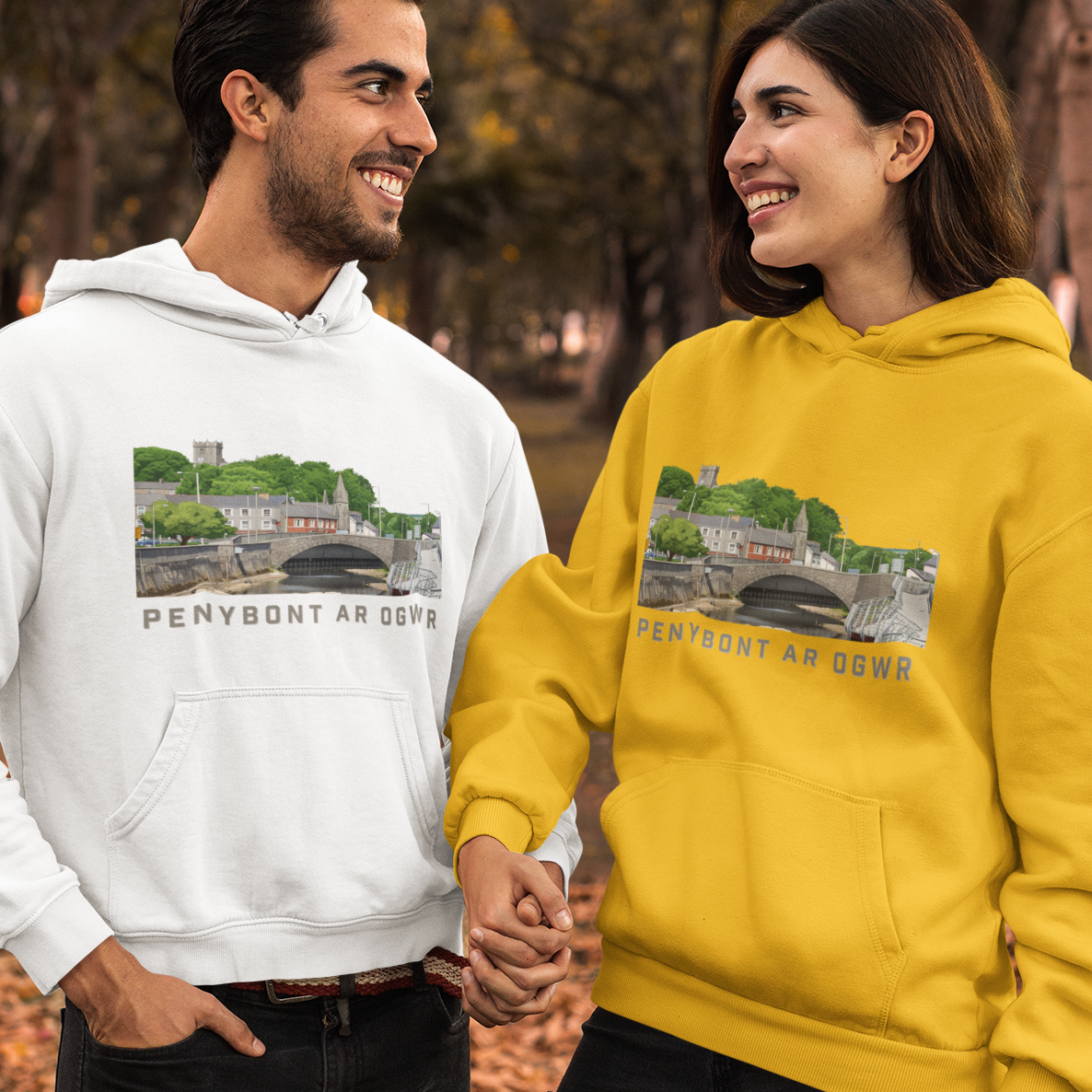 A man and a woman standing hand-in-hand in a park, wearing matching hoodies. The man wears a white hoodie and the woman wears a bright yellow hoodie, both featuring a graphic print of Penybont Ar Gofre and the text 'PENYBONT AR OGWR'.