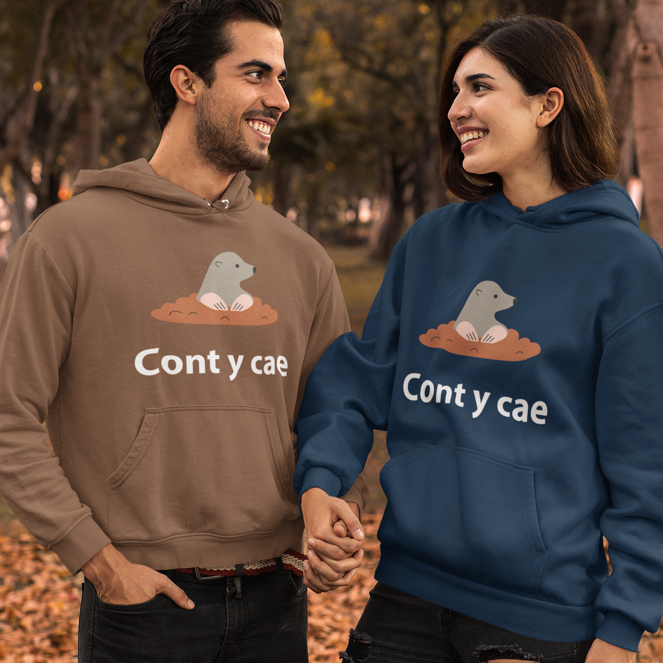 A man and a woman standing outdoors in a park holding hands, wearing matching hoodies with a cartoon frog graphic and the text 'Cont y cae'. The man's hoodie is brown and the woman's hoodie is navy blue.