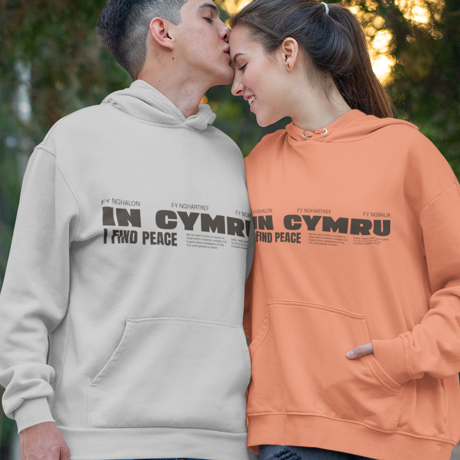 A man and a woman standing outdoors in front of trees, wearing matching pullover hoodies. The man wears a light beige hoodie with black text reading 'IN CYMRU I FIND PEACE', and the woman wears an orange hoodie with the same black text.