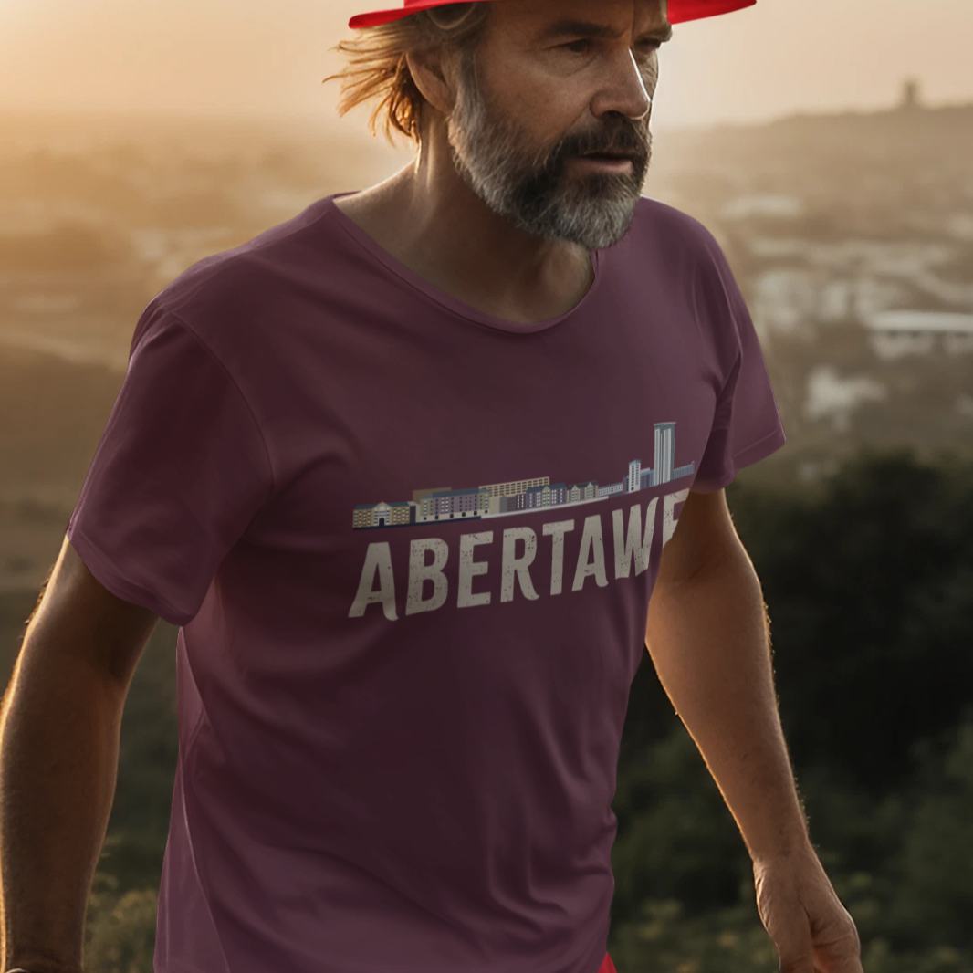 A man wearing a red bucket hat  and a mauve-purple t-shirt with 'ABERTAWE' printed on the front, walking outdoors against a warm sunset background.