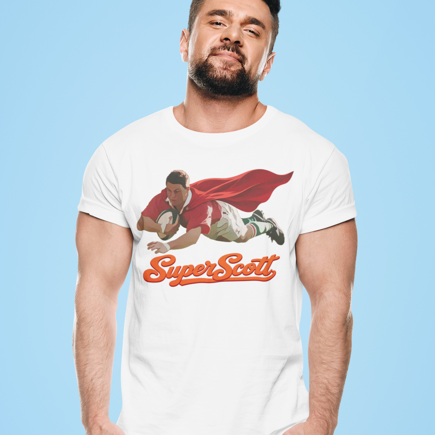 A man modeling a white graphic t-shirt featuring a cartoon illustration of a rugby player in a red cape with the text 'Super Scott' in orange. He is wearing blue sweatpants against a solid light blue background.