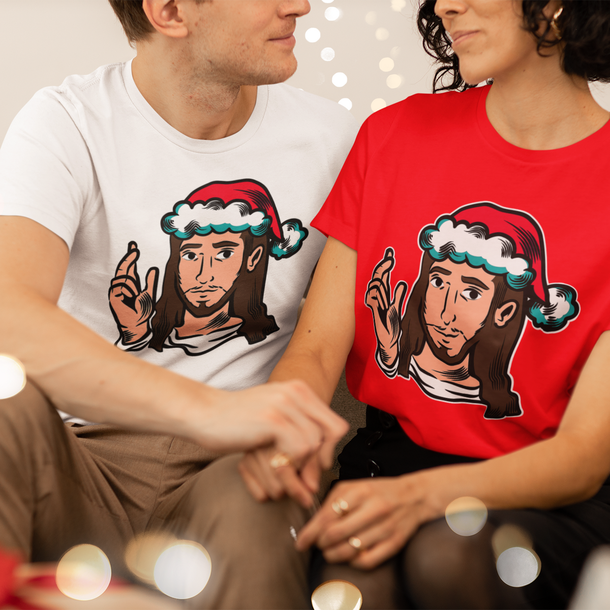 A man and a woman sitting closely together wearing matching graphic t-shirts with a cartoon Jesus design in a Santa hat. The man wears a white t-shirt and the woman wears a red t-shirt, with festive bokeh lights in the background.