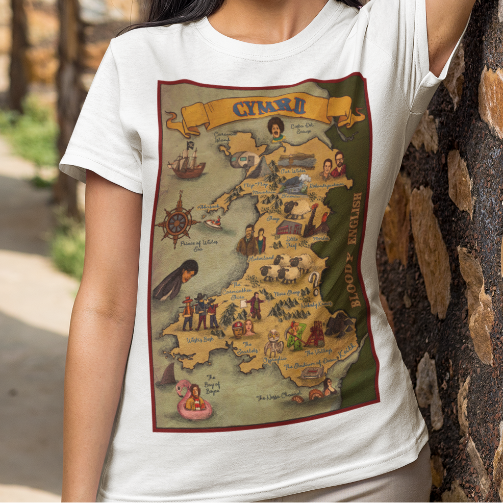 A woman posing outdoors against a stone wall, wearing a white crewneck t-shirt with a vintage-style map graphic of Wales on the front, paired with beige trousers.