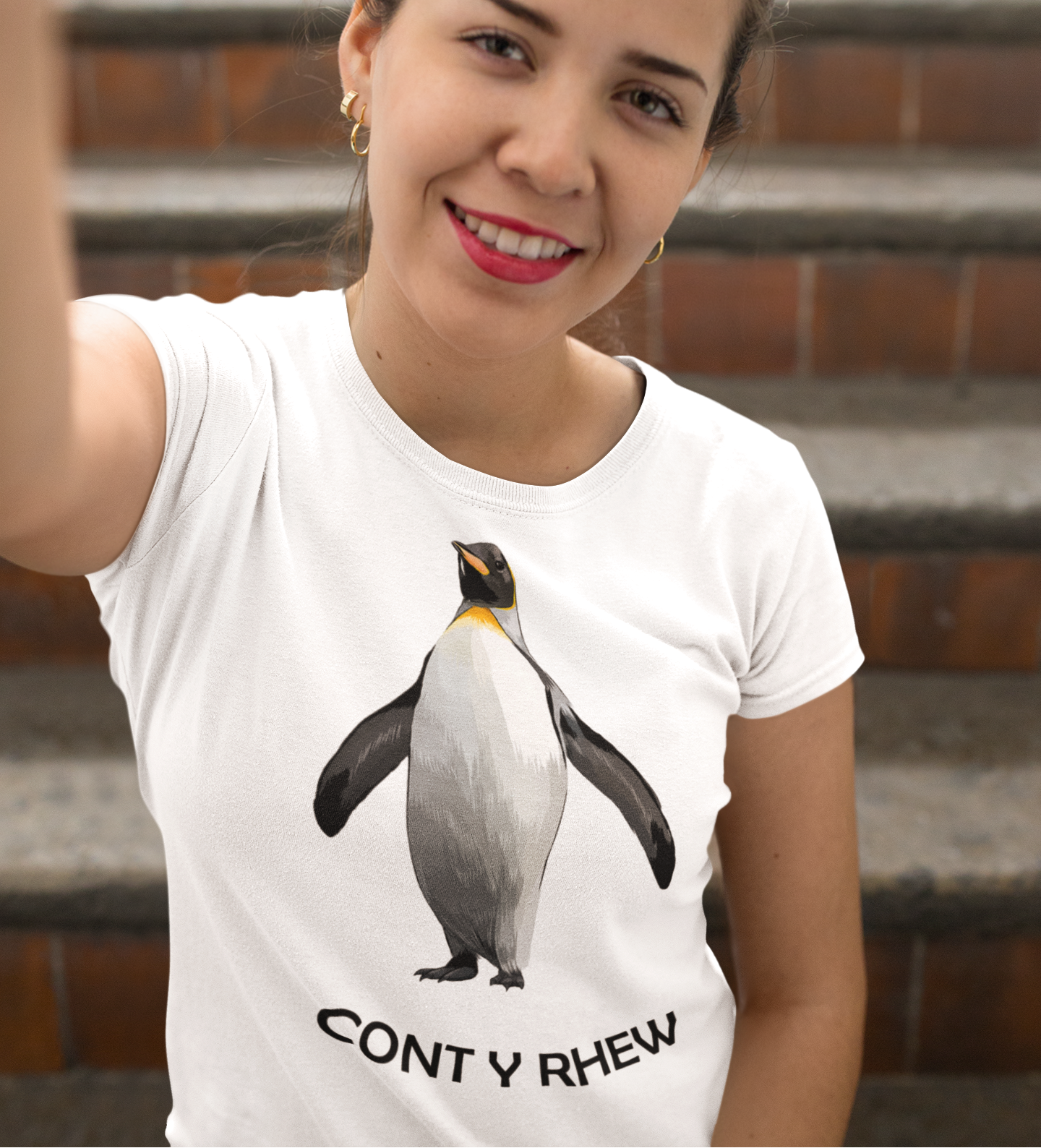 A woman wearing a white crewneck t-shirt with a graphic of a penguin and the Welsh text 'CONT Y RHEW' below it, posing in front of outdoor brick steps.