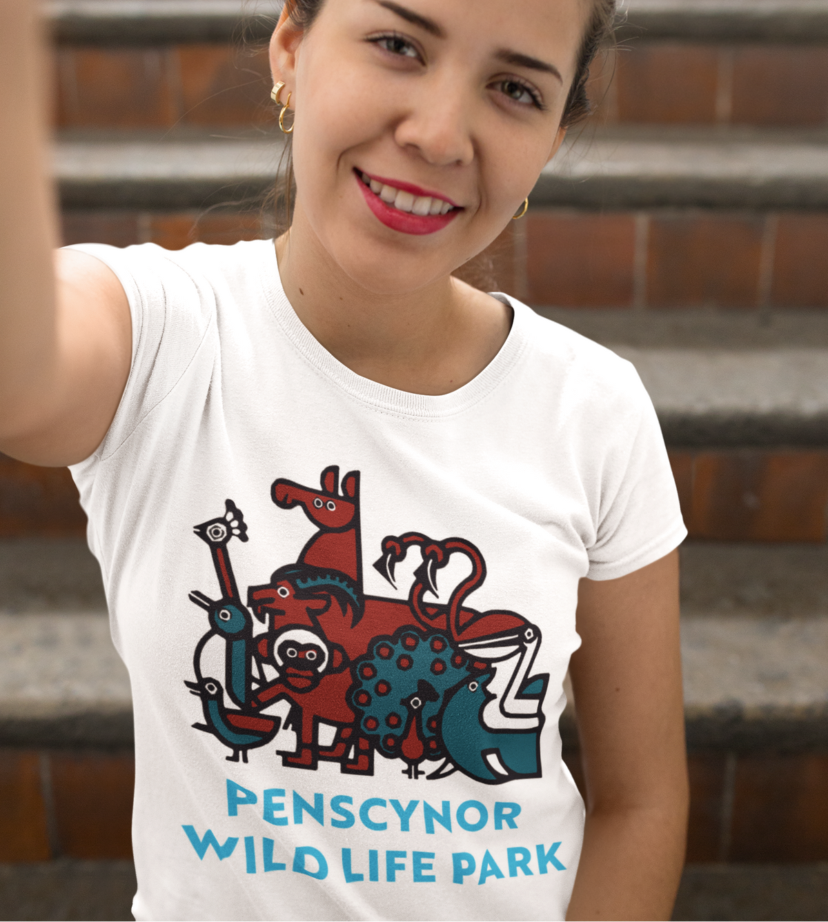 A woman wearing a white t-shirt with a colorful cartoon graphic of animals and the text 'PENSCYNOR WILDLIFE PARK', posing in front of outdoor brick steps.