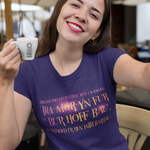 A woman taking a selfie in a cafe, wearing a purple t-shirt with a text print in pink and orange, while holding a white ceramic mug.