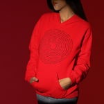 A model wearing a bright red hoodie with a black circular concentric text graphic on the front, paired with black pants, set against a solid dark red background.
