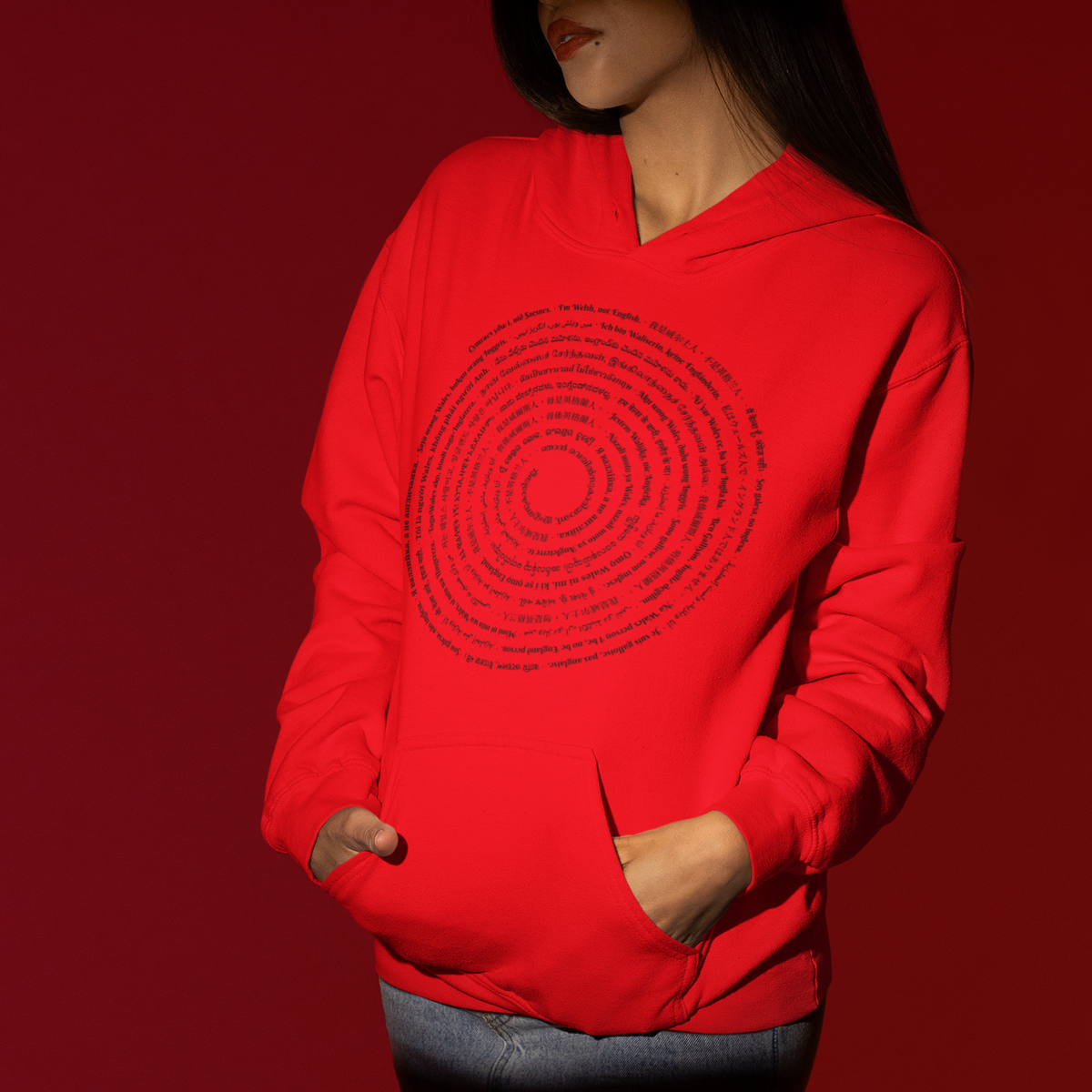 A model wearing a bright red hoodie with a black circular concentric text graphic on the front, paired with black pants, set against a solid dark red background.