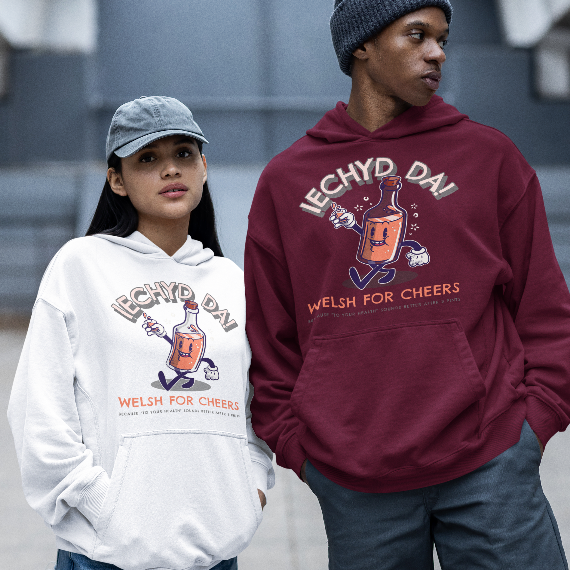 Two models wearing matching graphic hoodies in white and maroon, standing outdoors on a concrete walkway. The hoodies feature a cartoon mule graphic and the text 'IECHYD DA' and 'WELSH FOR CHEERS'.
