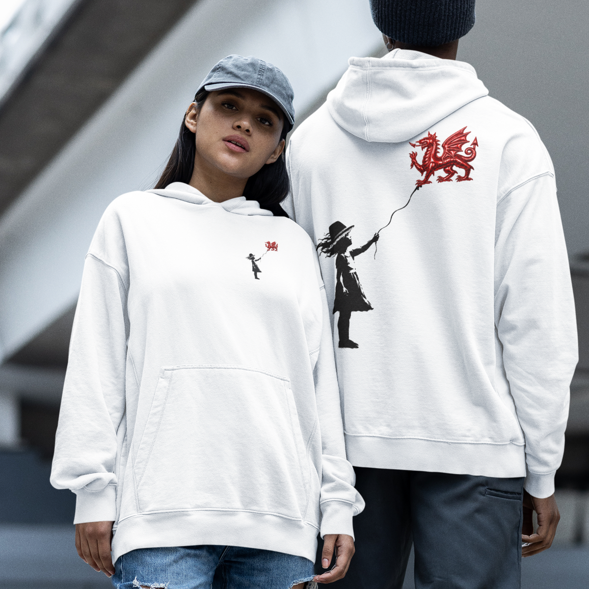 Two models wearing white hoodies in an outdoor urban setting. The model on the left wears a white hoodie with a small red rose graphic on the chest, paired with ripped blue jeans, a gray beanie, and red sneakers.
