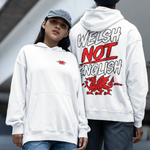 Two models standing outdoors in front of a modern concrete structure, showcasing the Welsh not English white hoodie. The model on the left wears a white hoodie with a small red dragon on the chest, paired with ripped blue jeans and red sneakers.