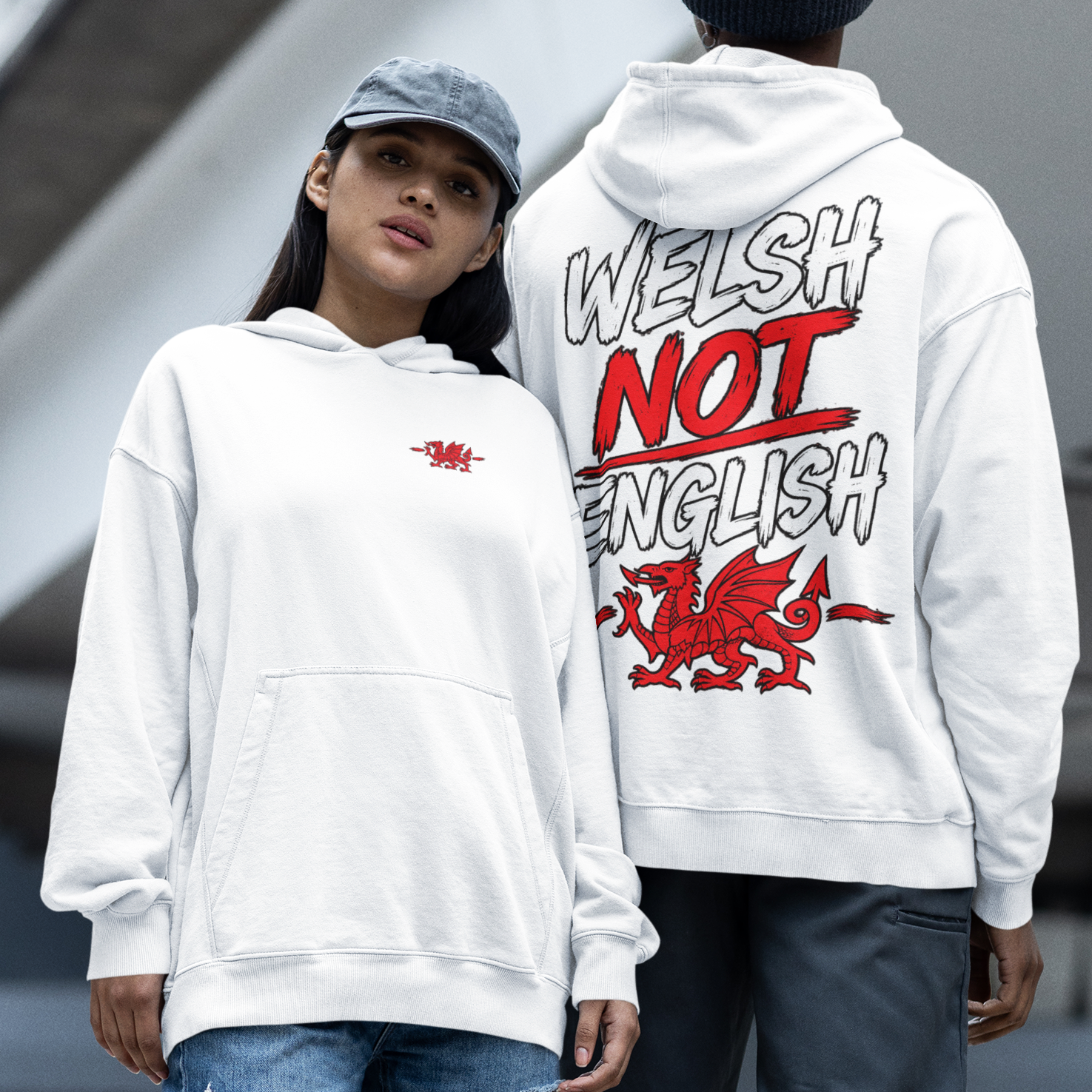Two models standing outdoors in front of a modern concrete structure, showcasing the Welsh not English white hoodie. The model on the left wears a white hoodie with a small red dragon on the chest, paired with ripped blue jeans and red sneakers.
