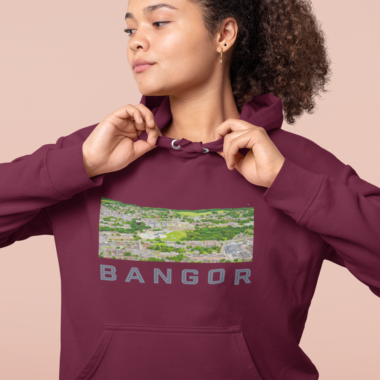 A woman wearing a maroon pullover hoodie with a rectangular scenic graphic on the chest and the word 'BANGOR' printed below it, posed against a solid light peach background.
