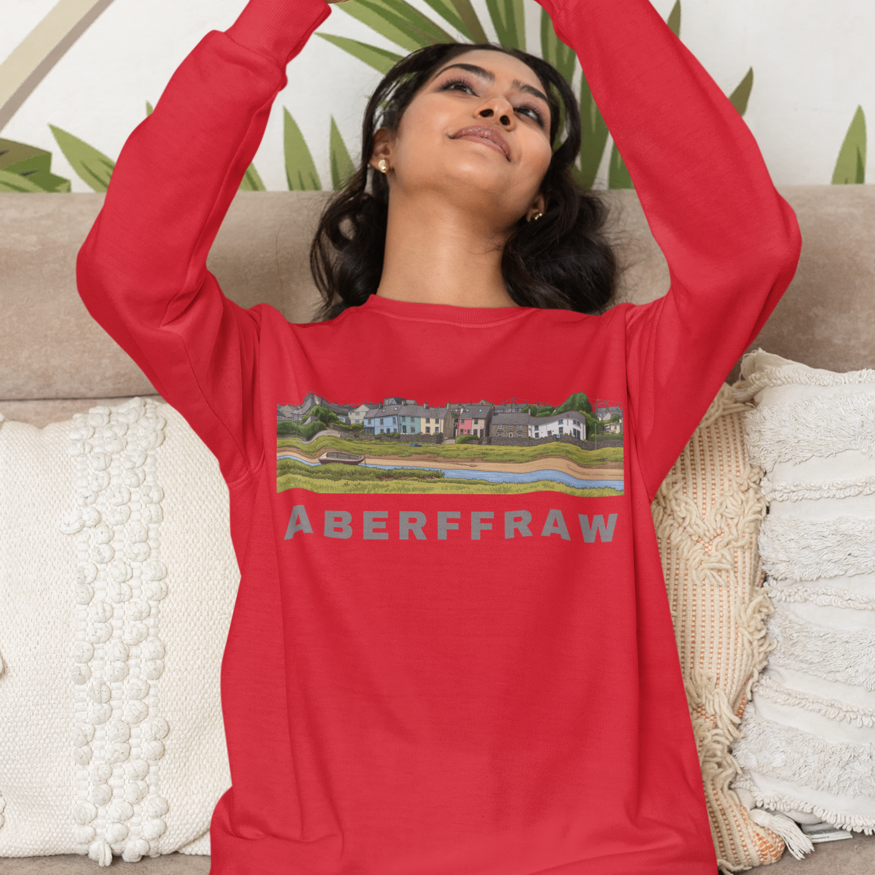 Person wearing a red sweatshirt with 'Aberffraw' text and graphic, sitting on a couch with a decorative plant in the background.