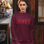 A woman wearing a burgundy crewneck sweatshirt with 'JONES' printed in red across the chest, paired with distressed light blue jeans, standing in front of a dark wooden storefront.