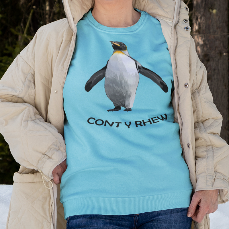 A person standing in the snow wearing a light blue crewneck sweatshirt with a penguin graphic and the text 'CONT Y RHEW', layered under a beige quilted puffer jacket, blue jeans, and black winter boots.