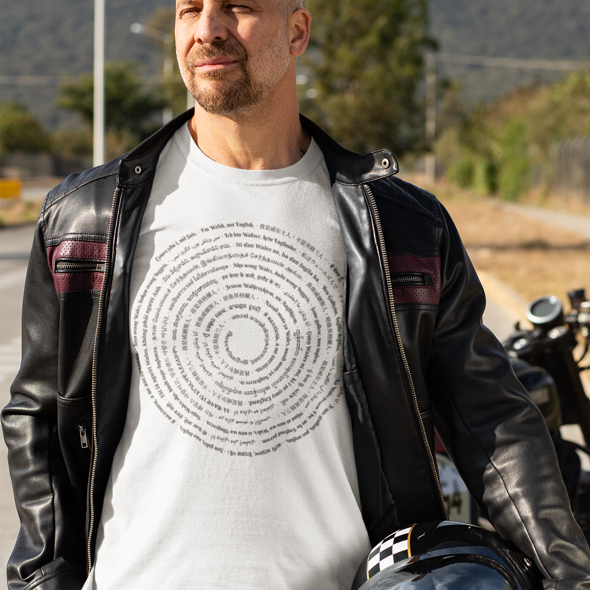 A man wearing a black leather jacket over a white graphic t-shirt and blue jeans stands on a paved road, holding a black motorcycle helmet. A motorcycle is parked in the background with hills and trees behind it.
