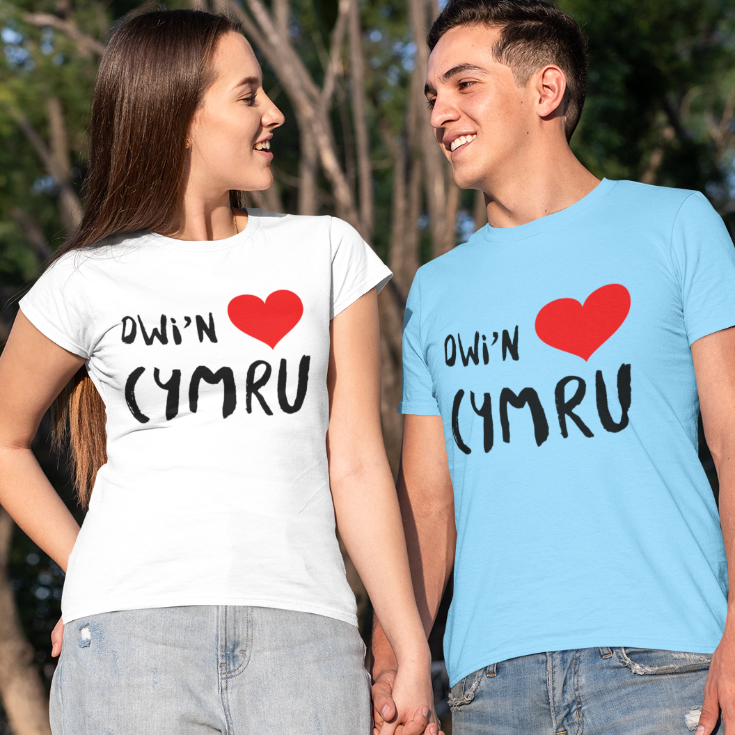 A man and a woman standing outdoors and holding hands, wearing matching graphic t-shirts with the Welsh phrase 'Dwl'n Caru Cymru' in black text and a red heart, one shirt being white and the other light blue.