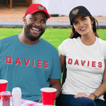 A man and a woman sitting outdoors on a lawn at a table, wearing matching t-shirts and caps with the name 'DAVIES' printed across them. The man wears a red cap and a teal t-shirt, while the woman wears a black cap and a white t-shirt.
