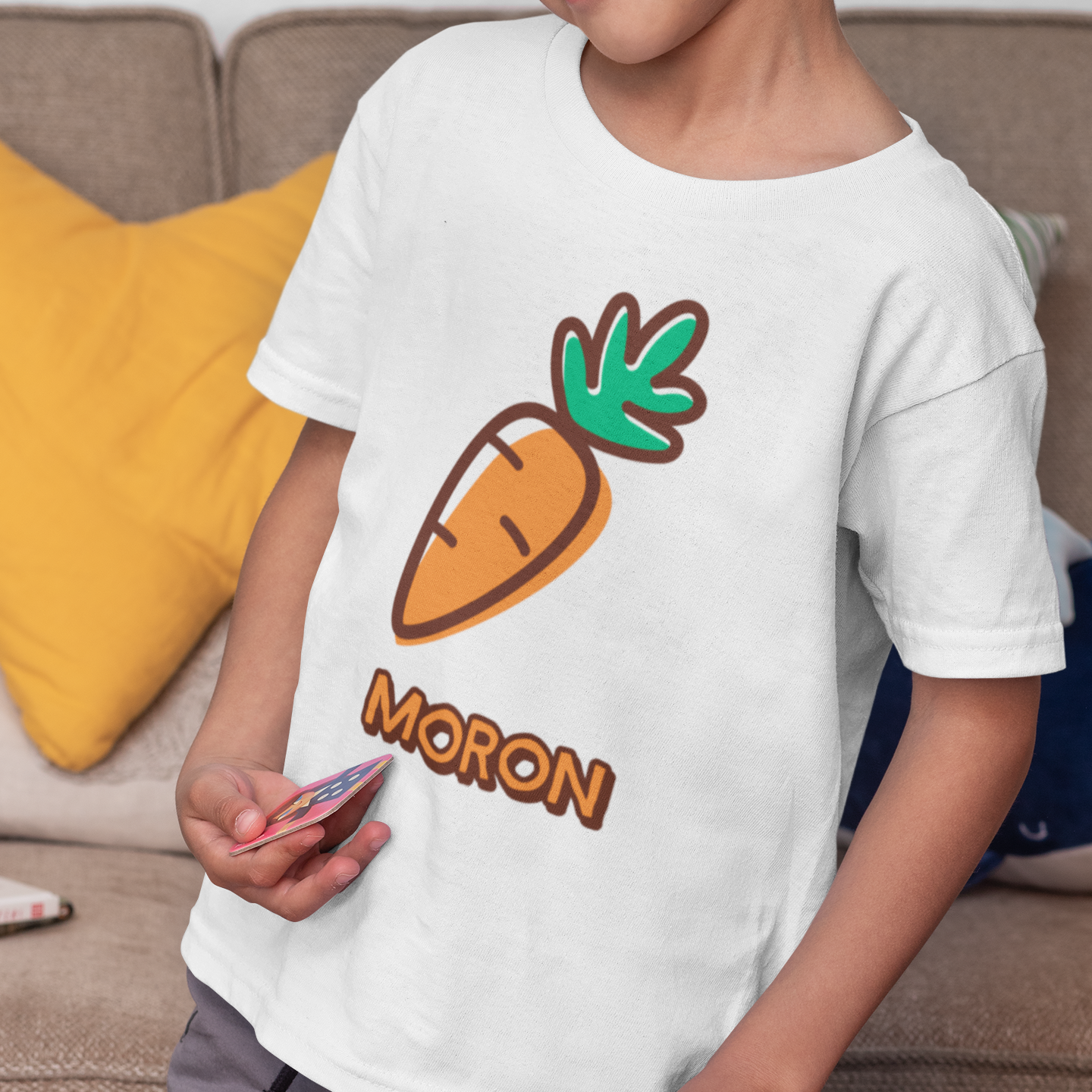 A young boy wearing a white t-shirt with a cartoon carrot graphic and the word 'MORON' printed below it, standing in a living room setting.