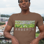 A man wearing a brown t-shirt with a green and white Arberth graphic print, standing on a balcony with a cityscape background.
