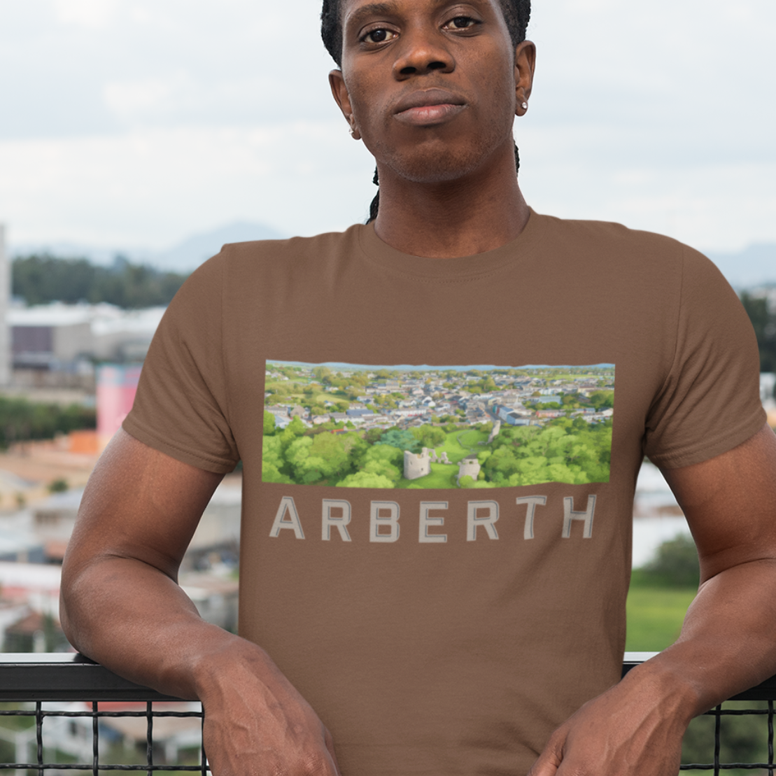 A man wearing a brown t-shirt with a green and white Arberth graphic print, standing on a balcony with a cityscape background.