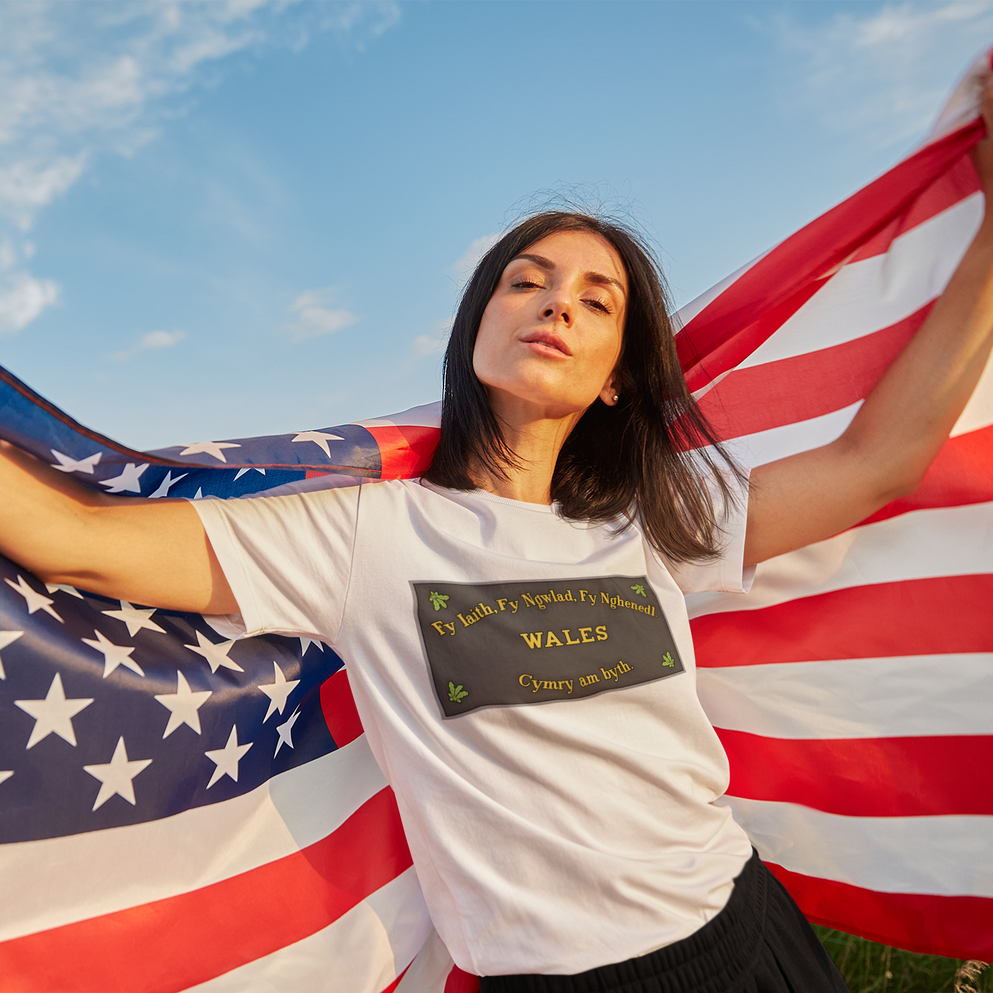 A woman outdoors holding a large American flag behind her, wearing a white T-shirt with a dark rectangular plaque from the Washingtom Monument.