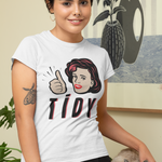 A woman wearing a white graphic t-shirt with a retro-style illustration of a woman giving a thumbs-up and the word 'TIDY' in black and red. She is standing indoors next to a gray tabby cat on a wooden table, with a framed art print on the wall behind her.