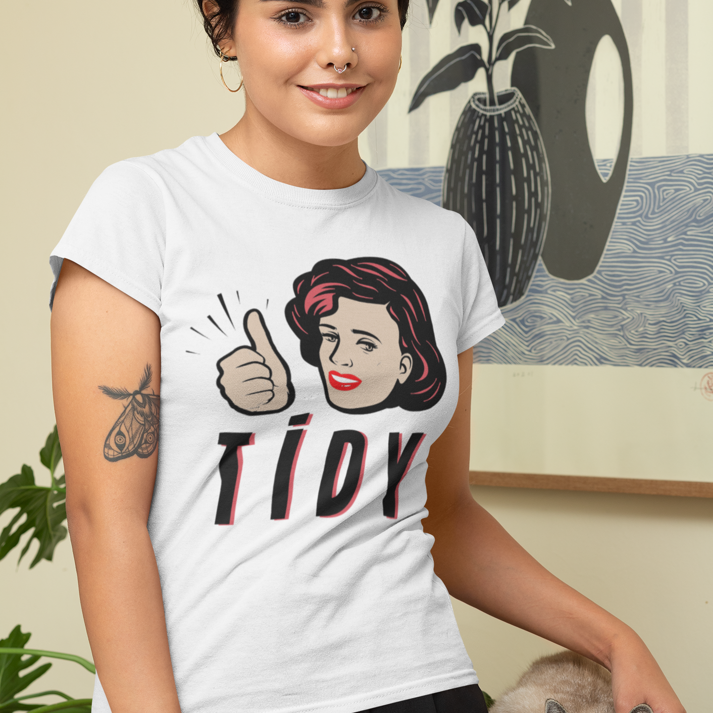 A woman wearing a white graphic t-shirt with a retro-style illustration of a woman giving a thumbs-up and the word 'TIDY' in black and red. She is standing indoors next to a gray tabby cat on a wooden table, with a framed art print on the wall behind her.